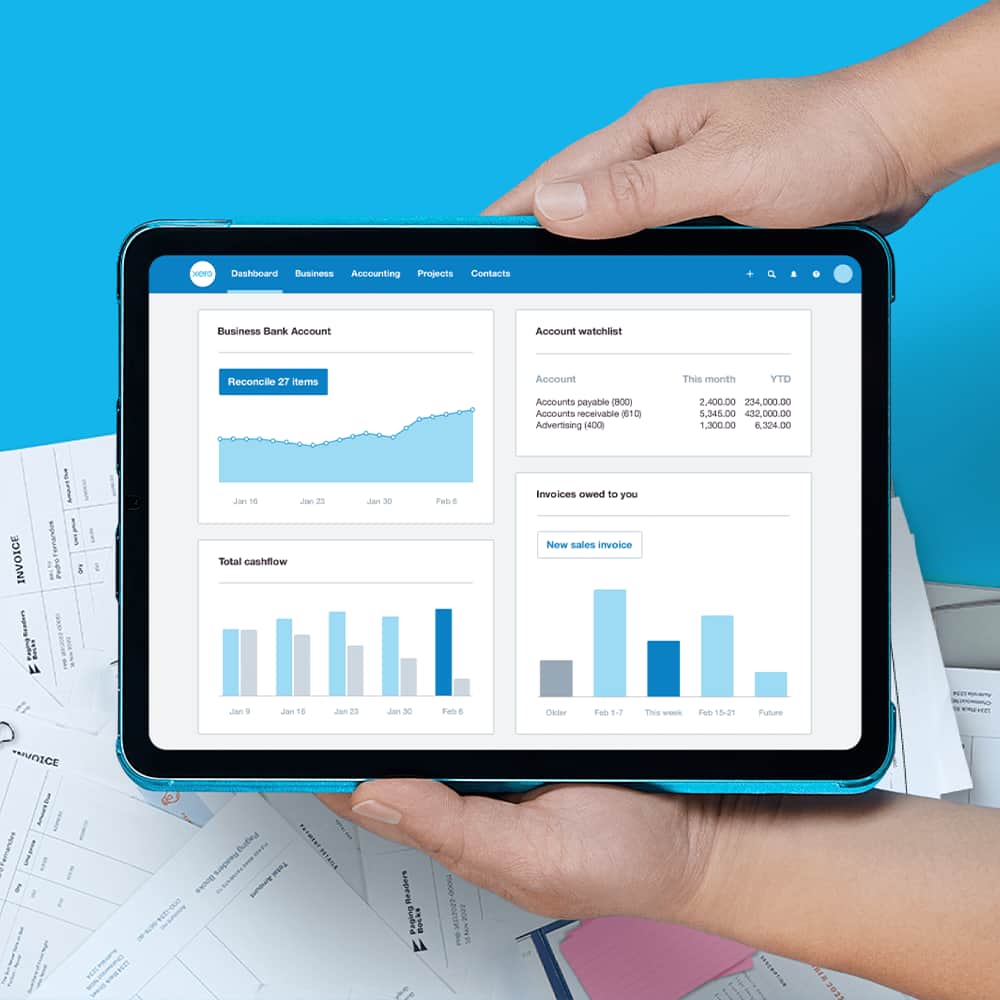 The Xero dashboard open on an iPad and held above scattered paper documents.